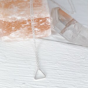 Puede incluir: Un collar de cadena de plata con un delicado colgante triangular. El collar se muestra sobre una superficie de madera blanca con una roca de sal rosa y un cristal de cuarzo transparente.