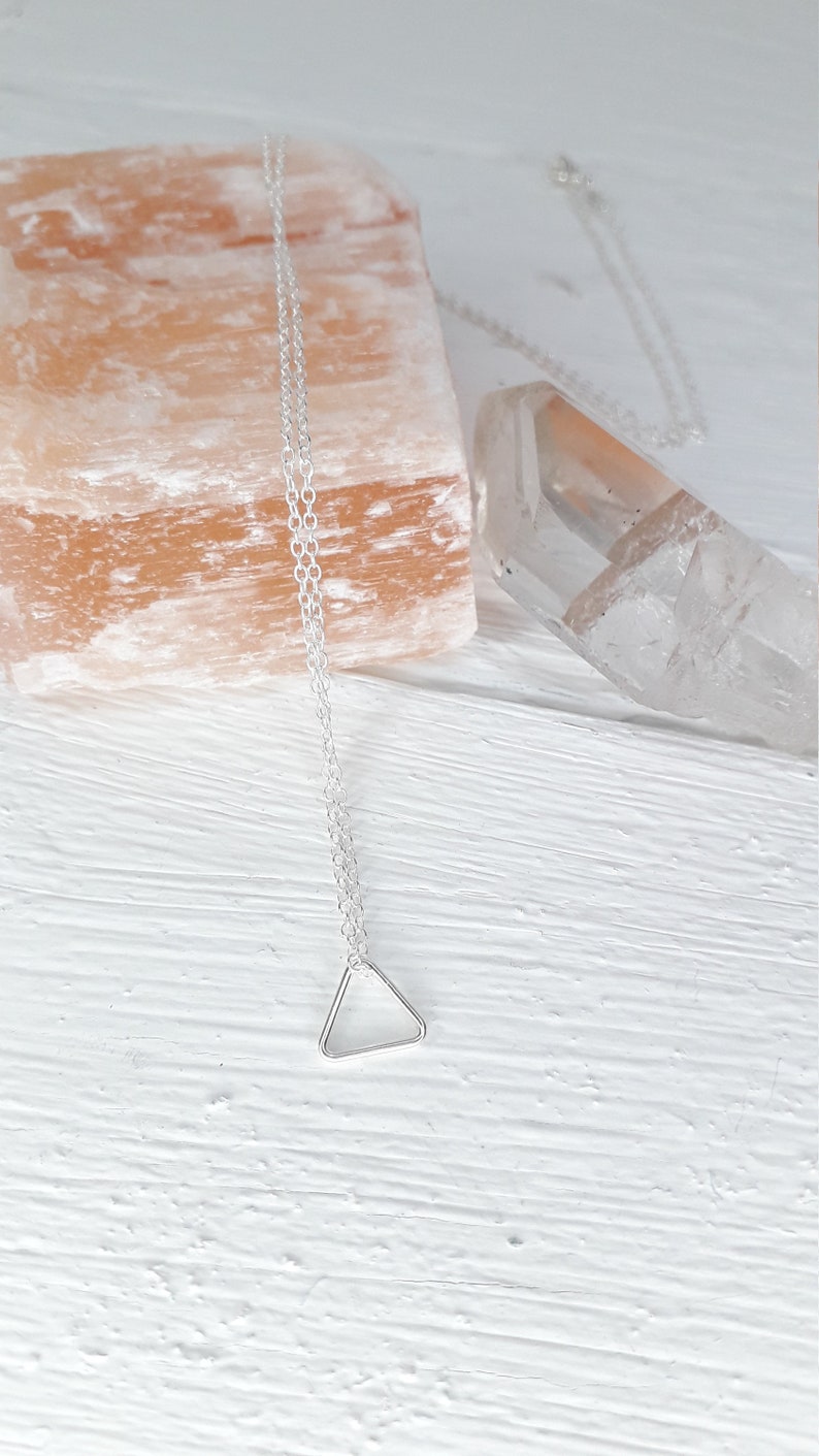 Puede incluir: Un collar de cadena de plata con un delicado colgante triangular. El collar se muestra sobre una superficie de madera blanca con una roca de sal rosa y un cristal de cuarzo transparente.