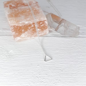 Puede incluir: Un collar de plata con un colgante triangular en una cadena delicada. El collar se muestra sobre una superficie de madera blanca con una roca de sal rosa del Himalaya y un cristal de cuarzo transparente.