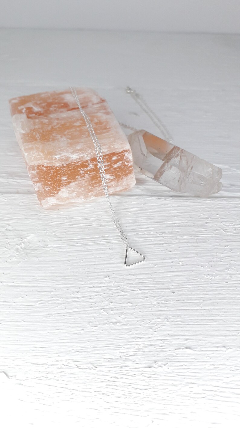 Puede incluir: Un collar de plata con un colgante triangular en una cadena delicada. El collar se muestra sobre una superficie de madera blanca con una roca de sal rosa del Himalaya y un cristal de cuarzo transparente.