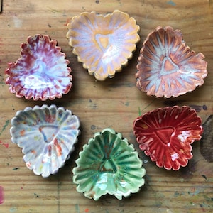 May include: Six handmade ceramic leaf-shaped dishes in various colours. The dishes are arranged in a circle and feature a variety of glazes, including red, orange, green, and blue. Each dish has a unique pattern and texture, ideal for serving snacks or as decorative pieces.