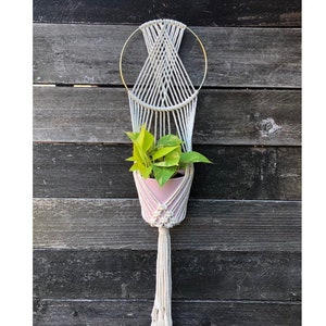 May include: A white macrame plant hanger with a gold metal ring at the top. A pink planter with a green plant is hanging in the macrame.