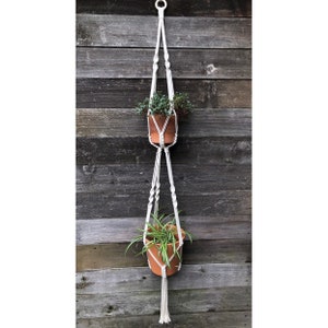 May include: A white macrame plant hanger with two hanging pots. The hanger is made of cotton rope and has a wooden ring at the top. The pots are terracotta and contain green plants.