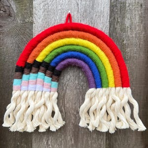 May include: A handmade rainbow wall hanging with a red top, transitioning through orange, yellow, green, blue, and purple. The bottom features cream-colored fringe. The rainbow is mounted on a wooden background.