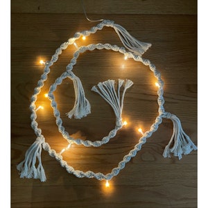 May include: A white cotton rope garland with small white tassels and warm white LED lights. The garland is coiled in a circle on a wooden surface.