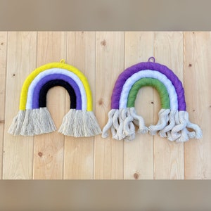 May include: Two rainbow-colored macrame wall hangings. The first is yellow, white, black, purple, and yellow. The second is purple, white, green, and white. Both have white fringe.