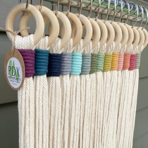 May include: A macrame plant hanger with wooden rings and a variety of colorful wrapped sections. The plant hanger features a cream-colored rope with sections wrapped in shades of purple, teal, navy, gray, and other colors. A tag with the text "PDX HOUSEPLANTS Handmade Macrame" is attached.