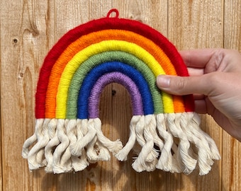 May include: A handmade rainbow wall hanging with a red, orange, yellow, green, blue, and purple design. The rainbow is made of yarn and has a macrame fringe at the bottom. The wall hanging is approximately 8 inches wide.