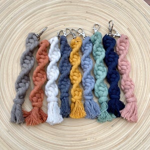 May include: Eight colorful macrame keychains with tassels. The keychains are made of twisted rope in shades of gray, orange, white, blue, yellow, light gray, green, and pink.