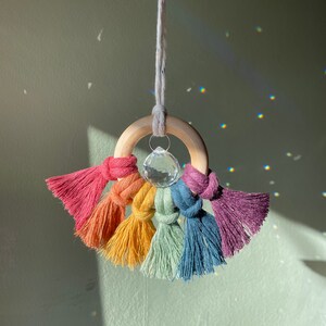May include: A rainbow macrame wall hanging with a wooden ring and a crystal pendant. The tassels are in red, orange, yellow, green, blue, and purple. The crystal reflects light, creating a sparkling effect. The wooden ring has a diameter of approximately 7 cm.
