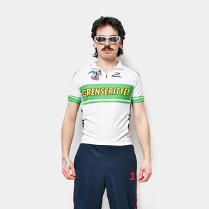 Op de afbeelding: Een wit fietsshirt met korte mouwen, groene en rode details en de tekst "GRENSERITTET" op de borst. Gecombineerd met een marineblauwe trainingsbroek met rode strepen. Vintage outfit.