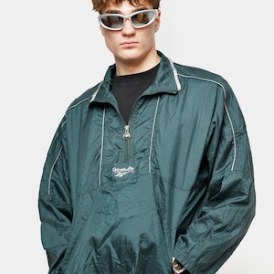 Op de afbeelding: Een donkergroene Reebok pulloverjas met een halve ritssluiting en witte bies. De jas heeft een relaxte pasvorm en het Reebok-logo op de borst. Het model draagt een zonnebril en een blauwe korte broek.