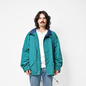 Peut inclure: Un coupe-vent sarcelle avec un col bleu foncé, porté sur un t-shirt blanc et un jean bleu clair. La veste présente une fermeture éclair frontale avec des boutons-pression en métal, des manches longues à poignets élastiques et un cordon.