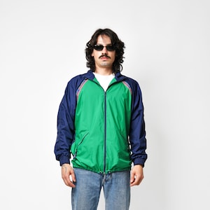 May include: A zip-up windbreaker jacket featuring a bright green centre panel, dark blue sleeves, and a hood. The jacket includes a white undershirt, blue denim jeans, and white athletic trainers with black accents. The jacket has a patterned trim.