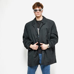 May include: A black overcoat with a button closure, worn over a black t-shirt and blue jeans. The person is wearing white sneakers with blue accents and silver-framed sunglasses. A silver watch is visible on the wrist.