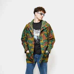 May include: A person in a camouflage jacket, black graphic t-shirt, blue jeans, and white trainers with blue detailing. The jacket has a green, brown, and tan pattern. The t-shirt features a graphic design. The person is wearing sunglasses.