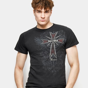 Może przedstawiać: Czarny t-shirt z czerwono-szarym wzorem krzyża z napisem "Hard Rock Cafe Munich". Koszulka ma krótkie rękawy i okrągły dekolt. Model ma na sobie niebieskie dżinsy.