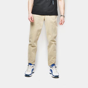 Puede incluir: Una persona vestida con una camiseta negra con gráficos, pantalones color beige y zapatillas blancas y azules. La camiseta presenta un diseño gráfico blanco y azul. Los pantalones son de color beige claro. Las zapatillas son blancas con detalles azules.