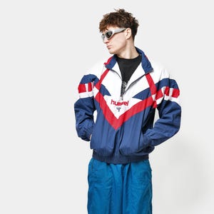 May include: A vintage blue, white, and red Hummel track suit. The jacket has a color block design with a zipper and the Hummel logo. The matching pants are blue with red and black accents. The outfit is paired with white and blue sneakers.