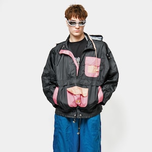 Könnte beinhalten: Eine schwarze und rosa Windjacke mit Kapuze, kombiniert mit einer blauen Trainingshose. Die Jacke hat rosa Taschen und Akzente. Die Trainingshose hat schwarze und rote Streifen an den Seiten. Weiße und blaue Turnschuhe vervollständigen das Outfit.