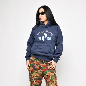 Può includere: Felpa con cappuccio blu navy con la scritta "PEAKPERFORMANCE 1986" e un logo bianco. L'outfit comprende pantaloni cargo mimetici nei toni del marrone, verde e rosso. La persona indossa occhiali da sole e scarpe da ginnastica.