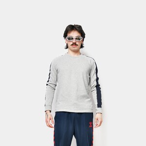 May include: A person wearing a grey long-sleeved top with navy stripes on the sleeves and navy track pants with red stripes. The person is also wearing black trainers and sunglasses.
