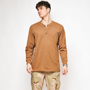 Könnte beinhalten: Ein langärmeliges braunes Henley-Shirt mit einer Knopfleiste mit drei Knöpfen und einer kleinen Brusttasche, kombiniert mit einer Camouflage-Cargohose mit beigem, braunem und sandfarbenem Muster sowie weißen Sportschuhen mit schwarzen und grauen Akzenten.