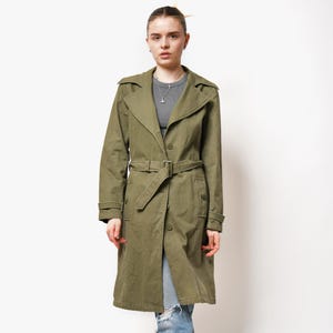 May include: A long, olive green trench coat with a belt and pockets. The coat is worn by a person with blonde hair and is standing on a white background.