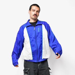 May include: A blue, white, and teal vintage windbreaker jacket with a full zipper. The jacket has a logo on the chest and teal trim. The model is wearing black cargo shorts and gray sneakers.