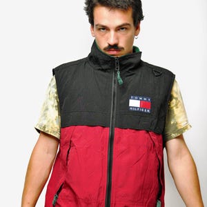 May include: A black and red Tommy Hilfiger vest with a zip-up front. The vest has a high collar and a small logo patch. The model is wearing a tie-dye shirt underneath and blue jeans.