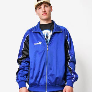 Könnte beinhalten: Eine blaue und schwarze Trainingsjacke mit weißen Paspeln und dem Logo "Jako" auf der linken Brust. Die Jacke hat einen Reißverschluss und einen Stehkragen.