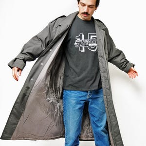 May include: A gray trench coat with a button closure, worn over a dark gray t-shirt and blue jeans. The t-shirt features the text "ROCK N ROLL MASKINE" and the number "15". The coat is open, revealing a light gray lining.