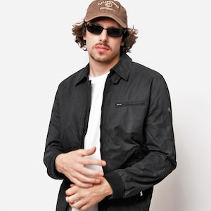 Puede incluir: Un hombre con una chaqueta negra, una camiseta blanca, pantalones de color caqui y una gorra de béisbol marrón con el texto "California University". Lleva gafas de sol.