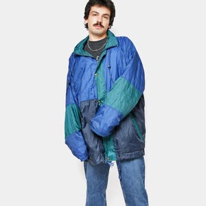 Może przedstawiać: Kurtka w stylu vintage w kolorach niebieskim i turkusowym. Kurtka ma wysoki kołnierz, sznurek i zapięcie na guziki. Osoba ma na sobie niebieskie dżinsy.