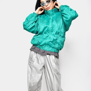 Peut inclure: Veste zippée vert turquoise avec col montant et pantalon de survêtement argenté. La veste a une texture froissée. Le pantalon est argenté métallisé avec des rayures latérales. La personne porte des lunettes de soleil argentées.