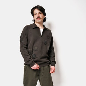 Puede incluir: Un hombre con una sudadera marrón oscuro con parches bordados negros y pantalones verde oscuro. Lleva zapatillas negras y blancas.