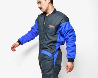 May include: A vintage blue and gray jumpsuit with the word "orion" in orange lettering on the chest. The jumpsuit has blue sleeves and accents, a front zipper, and is worn with silver sneakers.
