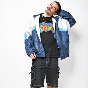 Op de afbeelding: Een persoon draagt een marineblauwe en witte windjack met capuchon, een zwart t-shirt met een kleurrijke afbeelding, een zwarte cargoshort, grijze sneakers en witte sokken. De windjack heeft een blauw, wit en lichtblauw kleurenschema.