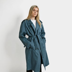 May include: A teal blue trench coat with a belt and pockets. The coat is worn by a person with long blonde hair.