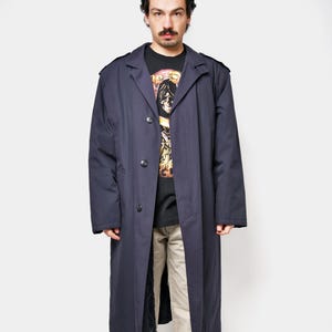 May include: A navy blue trench coat with a button-up front, worn over a black graphic t-shirt and khaki pants. The coat has shoulder epaulets and a long, flowing design. The person is wearing black and gray sneakers.