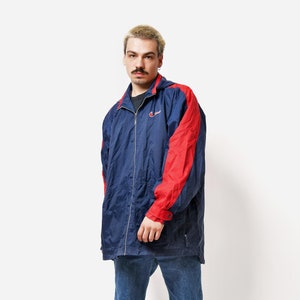 May include: A navy blue and red Champion jacket with a full zipper, paired with blue jeans and gray sneakers. The jacket has red accents on the sleeves and shoulders. The model is standing in front of a white background.