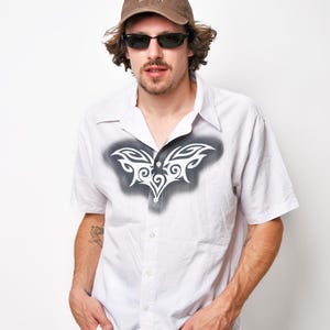 May include: A white button-down shirt with a tribal design on the front. The shirt has short sleeves and a collared neckline. The person is wearing a brown baseball cap with the text "CALIFORNIA UNIVERSITY" on it.