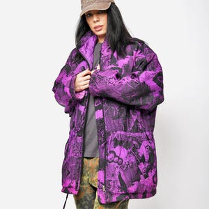 Peut inclure: Veste violette et noire à motifs avec une casquette marron. La veste a une fermeture éclair et des poches. La personne porte également un pantalon camouflage et un t-shirt gris. La casquette porte le mot "California".