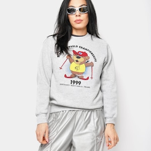 Peut inclure: Un sweat-shirt gris clair avec un dessin d'ours portant un chapeau rouge et des skis, avec le texte "WORLD CHAMPIONSHIPS 1999". Le sweat-shirt est associé à un pantalon de survêtement argenté.