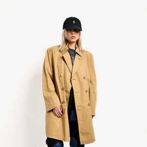 May include: A tan double-breasted trench coat with a black baseball cap. The person is wearing blue jeans and silver sneakers.
