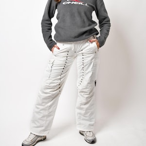 May include: A woman wearing a grey hooded sweatshirt with the O'Neill logo in red and white. She is also wearing white cargo trousers with silver studs and a navy blue baseball cap.