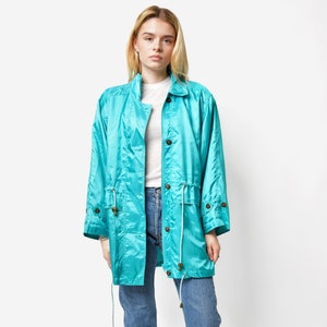 May include: A turquoise blue jacket with a drawstring waist and button closure. The jacket is made of a shiny, silky material. The model is wearing blue jeans and black sneakers.