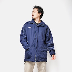 May include: A navy blue jacket with white trim and a zipper closure. The jacket has two front pockets and a small logo on the chest. The person is wearing khaki pants and black sneakers.