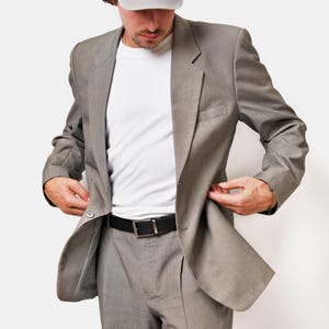 May include: A gray suit jacket with a white t-shirt underneath. The person is wearing a gray baseball cap.
