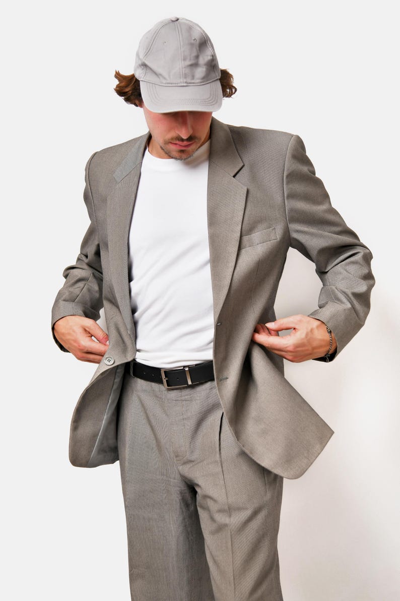 May include: A gray suit jacket with a white t-shirt underneath. The person is wearing a gray baseball cap.